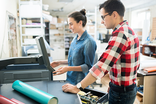 Smiling attractive young lady in casual shirt viewing information on monitor of printer while choosing print options with colleague in printing house