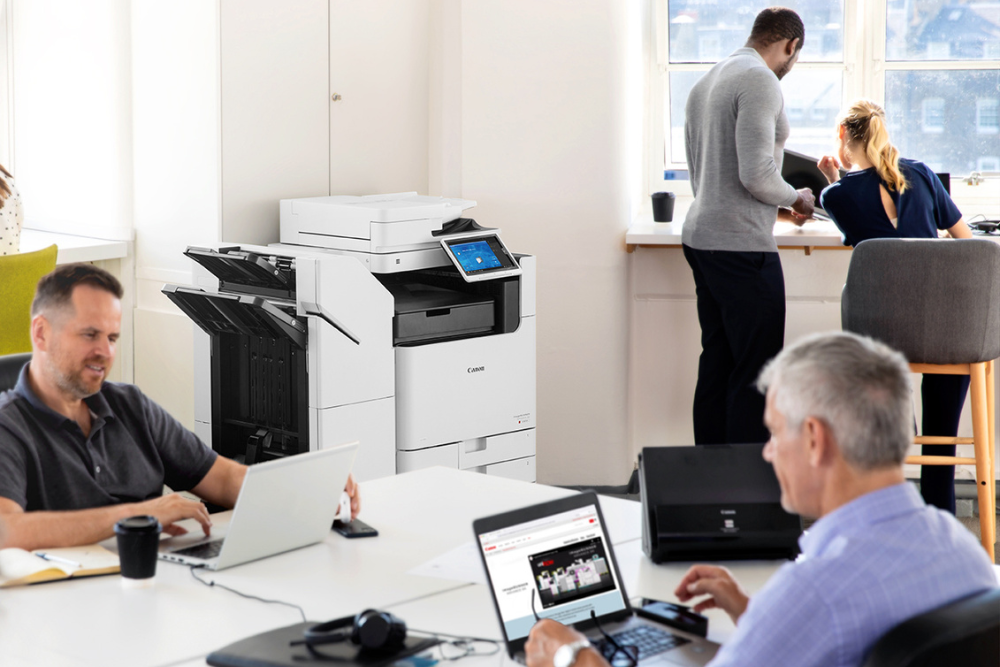 Office environment with employees working on laptops and a multifunction printer in the background. The multifunction printer is prominently displayed, showcasing its role in a busy office setting.