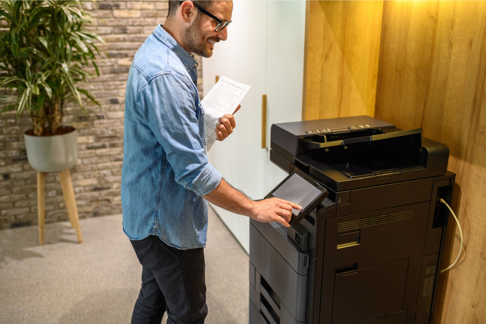 A man using an MFP (multifunction printer) in an office, interacting with the touchscreen while holding a document, showcasing the efficiency and versatility of MFP technology.