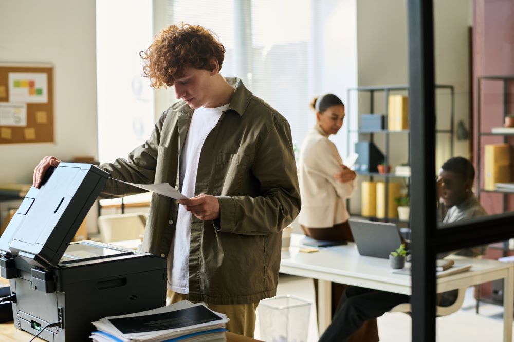 An office employee using a printer while colleagues work in the background, illustrating the efficiency of Managed Print Services in streamlining office printing tasks.