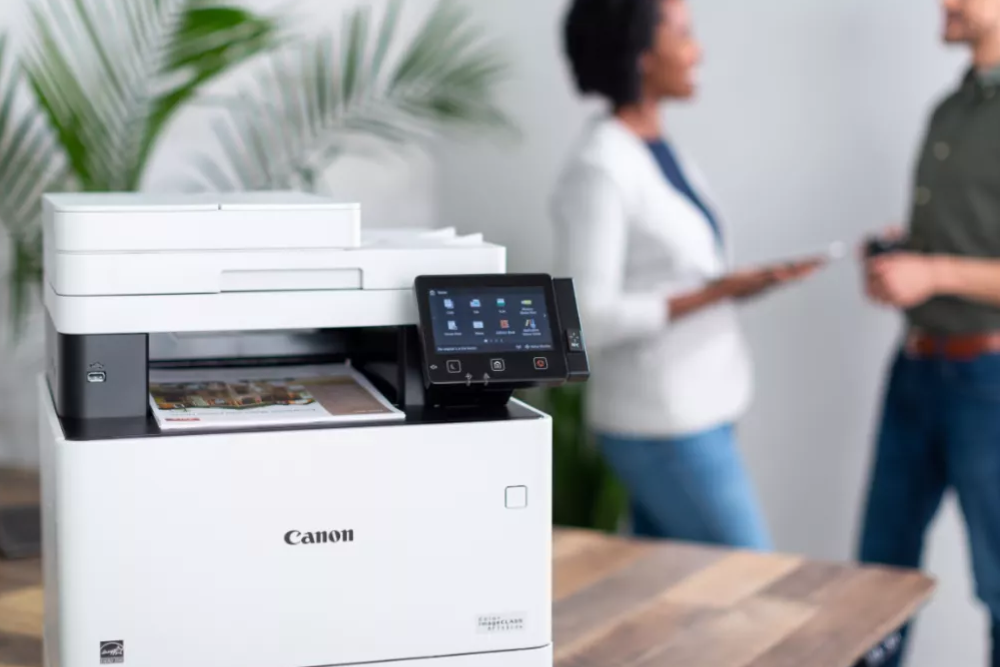 A sleek Canon copier placed on a wooden table in a modern office setting, with two professionals in the background having a discussion. The image highlights the functionality and reliability of Canon copiers for efficient document handling in a professional environment.